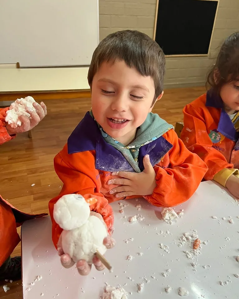 Un niño del jardín sonríe mientras modela una figura con masa en la mesa de trabajo