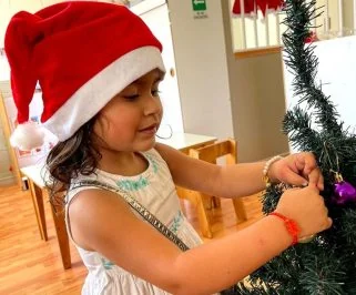 Una niña con gorro de Navidad cuelga un adorno en el árbol del jardín